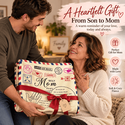 A man holding a wrapped fleece blanket for his mother in a cozy living room setting. The blanket features a vintage airmail design with red, white, and blue stripes, various postage stamp graphics, and a red ribbon with a kraft paper tag.