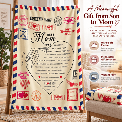 A personalized Mother's Day blanket displayed on a wooden easel in a cozy living room setting. The blanket features a cream background with red and blue striped borders resembling an airmail envelope, decorated with stamps, hearts, and a central heart-shaped lettering design that reads 'Best Mom ever'.