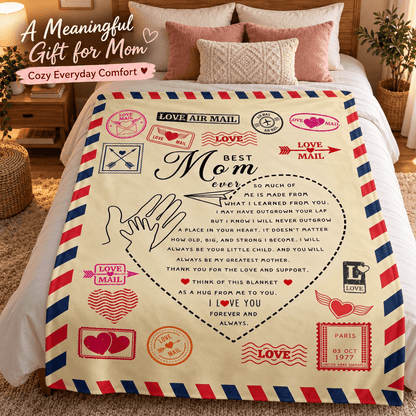 A cozy cream-colored blanket with a vintage airmail design and a heartfelt message for Mom, styled on a bed with pink pillows and a wooden headboard in a bedroom setting.