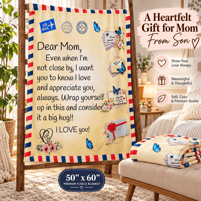 A fleece blanket with a vintage airmail design is draped over a wooden stand in a cozy living room setting. The blanket features a cream background with red and blue striped borders, blue butterflies, and a sentimental message for 'Mom'.
