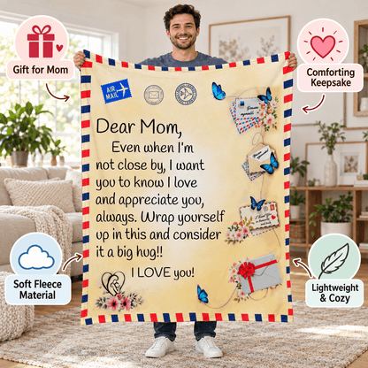 A man holds up a large fleece blanket with a printed sentimental message for Mom, styled in a bright living room. The blanket has an airmail-style red, white, and blue border, decorative icons, and text including 'Dear Mom, Even when I'm not close by, I want you to know I love and appreciate you, always.