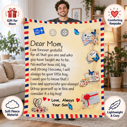 A man holds up a large fleece blanket with a printed heartfelt letter to Mom, styled like an airmail envelope with red, white, and blue borders and decorative graphics. The scene is set in a cozy living room, with circular callout badges around the edges highlighting gift, comforting keepsake, soft fleece material, and lightweight cozy qualities.
