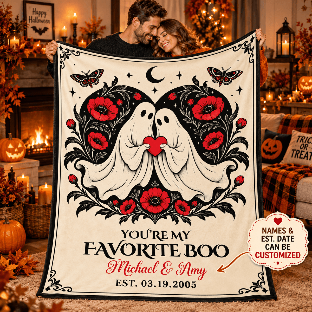 A couple holds up a large Halloween-themed fleece blanket in a cozy, decorated living room. The blanket features a cream background with a black and red graphic of two ghosts kissing, surrounded by poppy flowers, butterflies, and a crescent moon.