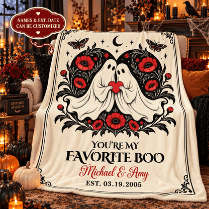 A personalized Halloween-themed blanket draped over a chair in a cozy, warmly lit living room. The blanket features a cream background with a black and red graphic of two ghosts surrounded by flowers, with the text 'YOU'RE MY FAVORITE BOO Michael & Amy EST.