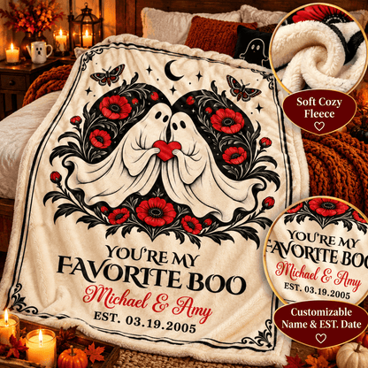 Personalized Halloween fleece blanket draped over a bed in a cozy bedroom setting. The blanket features a cream background with a black and white ghost design, red poppy flowers, butterflies, stars, and the text 'YOU'RE MY FAVORITE BOO Michael & Amy EST.
