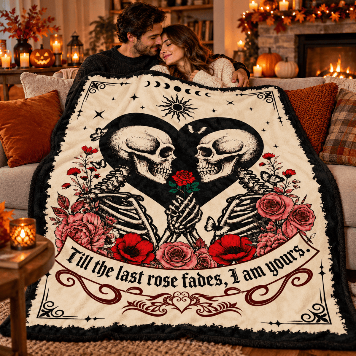 A couple sits on a couch in a cozy living room, wrapped together in a large cream-colored blanket featuring a black-and-white skeleton heart design, red roses, and the phrase 'Till the last rose fades, I am yours.'.