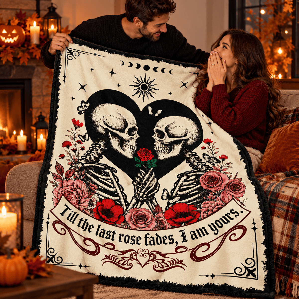 A couple holds up a cozy cream-colored blanket with a gothic skeleton design, featuring two skeletons holding a red rose between black hearts, surrounded by red roses and the text 'Till the last rose fades, I am yours.' The scene is set in a warmly lit living room decorated for Halloween.