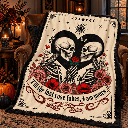 A Halloween-themed fleece throw blanket draped over a brown leather armchair in a cozy, warmly lit room. The blanket features a cream background with a black and white illustration of two skeletons forming a heart, surrounded by red and pink roses, stars, and a banner with gothic text.