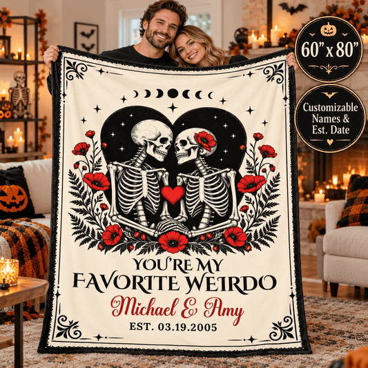 A couple holds up a large Halloween-themed fleece blanket in a cozy living room. The blanket features a cream background with a black border, two skeletons facing each other inside a black heart, red poppy flowers, and the text 'YOU'RE MY FAVORITE WEIRDO Michael & Amy EST.