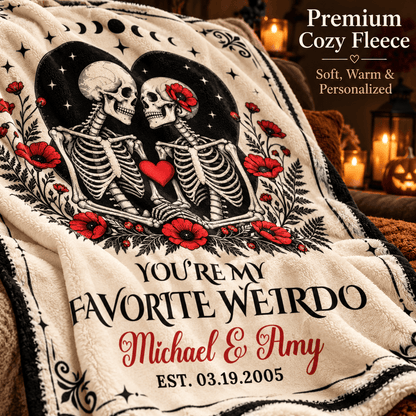 A personalized cream-colored fleece throw blanket draped over a couch, featuring a black-and-white skeleton illustration with red poppies, a red heart, and custom text reading 'YOU'RE MY FAVORITE WEIRDO Michael & Amy EST. 03.