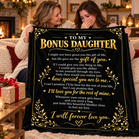 Two women sitting on a couch in a cozy living room setting, holding up a black blanket with a gold and white sentimental message addressed to a daughter.