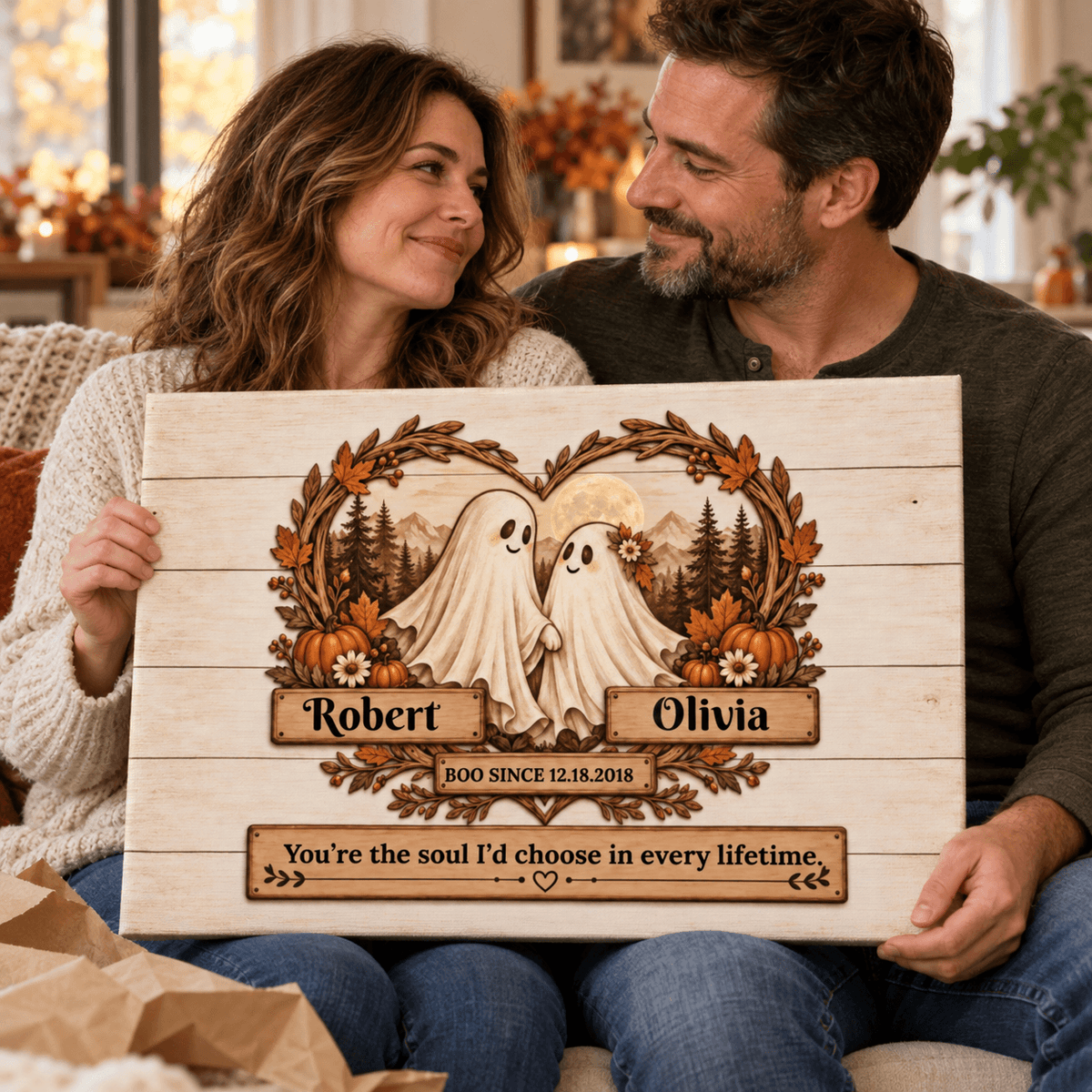 A couple sits on a couch holding a personalized wooden sign featuring two ghost figures framed by autumn leaves and pumpkins, with the names Robert and Olivia and a sentimental quote engraved on the wood.