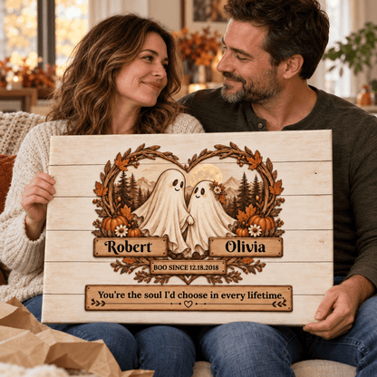 A couple sits on a couch holding a personalized wooden sign featuring two ghost figures framed by autumn leaves and pumpkins, with the names Robert and Olivia and a sentimental quote engraved on the wood.