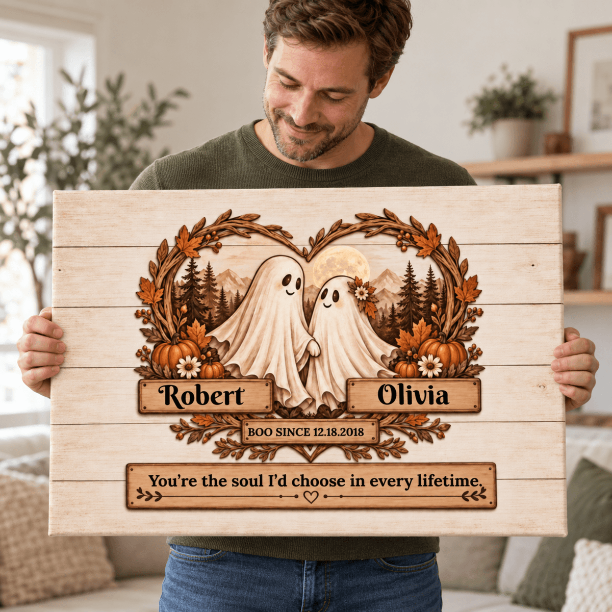 A man holds up a personalized wooden sign featuring a laser-burned design of a couple dressed as ghosts, framed by a leafy wreath and mountains. The names 'Robert' and 'Olivia' are printed on wooden plaques, with a bottom plaque reading 'You're the soul I'd choose in every lifetime.